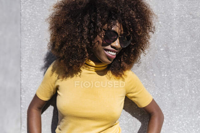 Giovane allegra femmina etnica con acconciatura afro ridere alla luce del sole vicino alla parete grigia — Foto stock