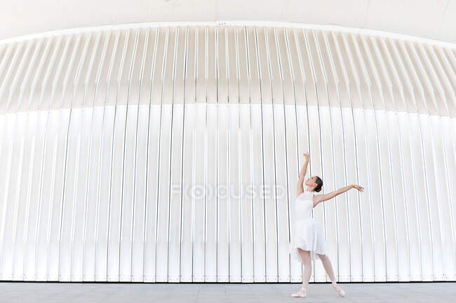 Вид сбоку на молодую балетную танцовщицу в пуантах с поднятыми ногами и руками, танцующую на тротуаре на открытом воздухе — стоковое фото