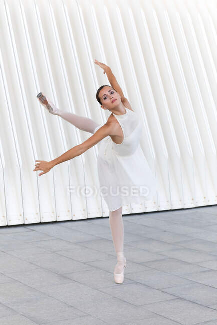 Ballerina di danza classica in punta di piedi in scarpe da punta con gamba rialzata e braccio che balla sul pavimento piastrellato all'aperto guardando la macchina fotografica — Foto stock