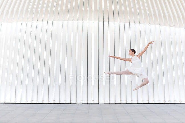 Young female ballet dancer in pointe shoes with raised leg and arm jumping over tiled pavement — Stock Photo