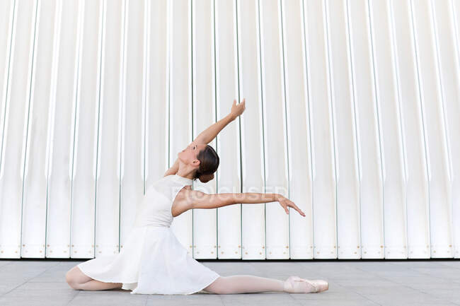 Vista lateral da jovem bailarina graciosa em vestido branco sentado na passarela durante a realização de pose de balé e olhando para cima — Fotografia de Stock