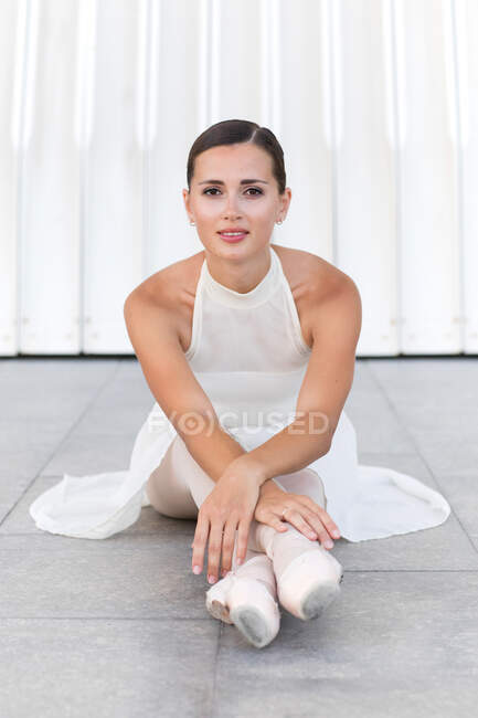 Joven encantadora bailarina de ballet en vestido blanco y zapatos puntiagudos mirando a la cámara mientras está sentada en la calle - foto de stock