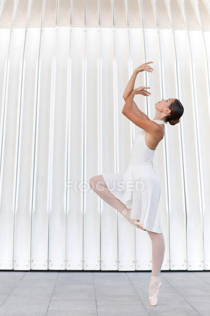 Side view of young female ballet dancer in tiptoe in pointe shoes with raised leg and arm dancing on tiled pavement outdoors — Stock Photo