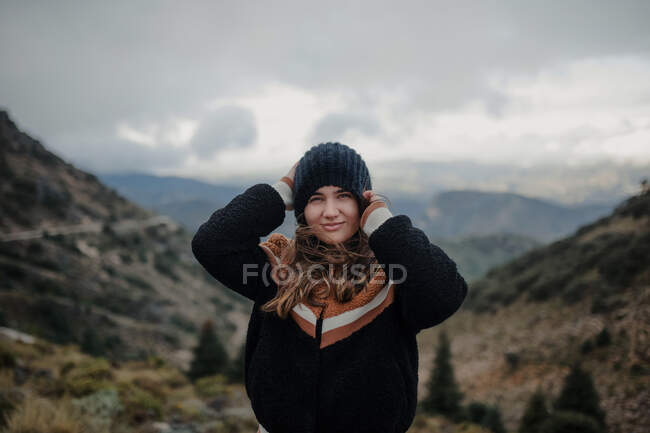 Sorrindo jovem fêmea colocando chapéu quente e olhando para a câmera enquanto estava em planaltos ásperos no dia sombrio nublado — Fotografia de Stock