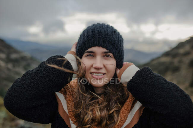 Smiling young female putting on warm hat and looking at camera while standing on rough highlands on cloudy gloomy day — Stock Photo