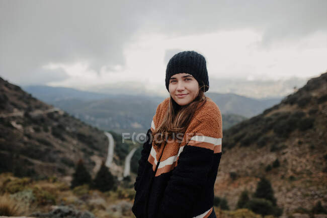 Jeune femme souriante regardant la caméra tout en se tenant debout sur les hautes terres rugueuses par temps nuageux sombre — Photo de stock