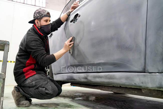 Vista lateral del maestro masculino en el automóvil de pulido de ropa de trabajo sucia mientras se prepara el vehículo para pintar en servicio de reparación - foto de stock