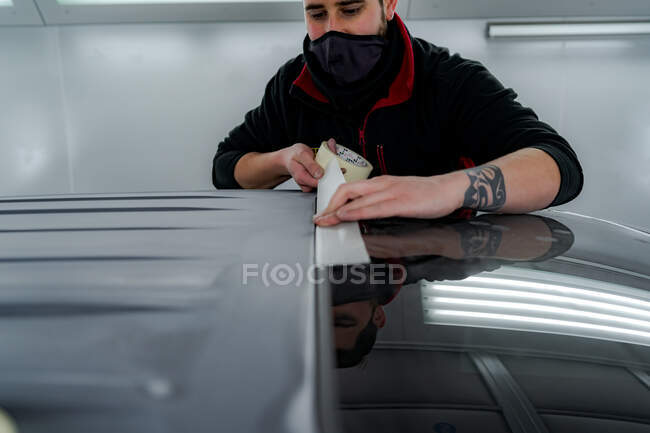 Busy male mechanic in mask sticking tape on car before painting in workshop — Stock Photo