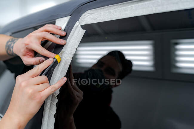 Чоловік-працівник наклеює захисну стрічку на автомобіль під час підготовки автомобіля для роботи з фарбою в майстерні при використанні канцелярського ножа — стокове фото
