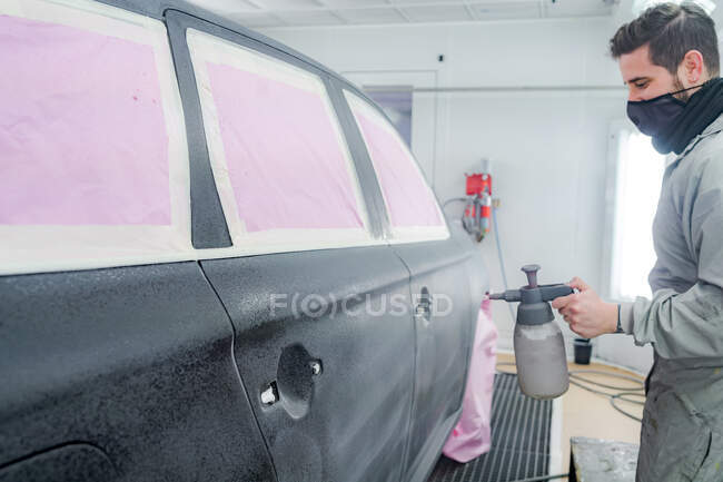 Side view of male worker in mask and uniform painting automobile while using paint gun — Stock Photo
