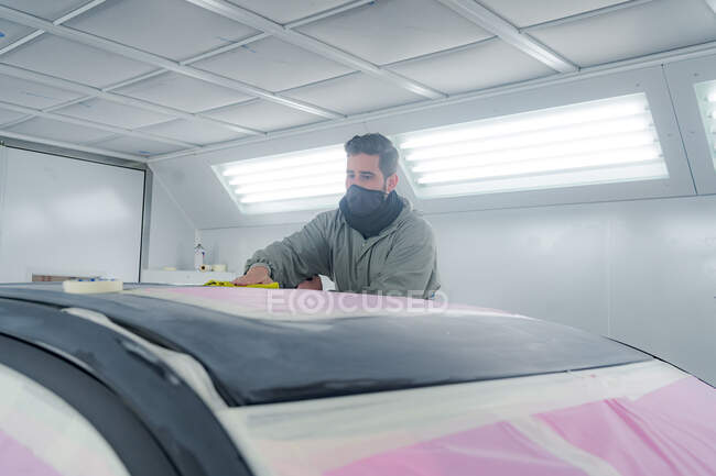 Männlicher Meister in schmutziger Arbeitskleidung poliert Automobil, während er Fahrzeug für die Lackierung im Reparaturservice vorbereitet — Stockfoto