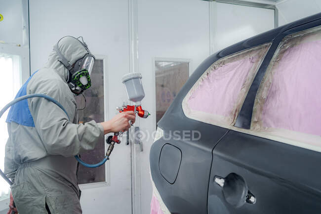 Vista lateral do macho na máscara respiratória e no carro protetor da pintura do terno com pistola de pulverização no serviço — Fotografia de Stock
