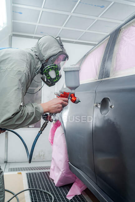 Вид збоку чоловіка в респіраторній масці і захисний костюм малювання автомобіля з розпилювачем на службі — стокове фото
