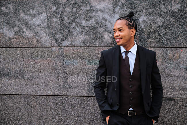 Decidido sonriente empresario afroamericano en traje elegante de pie con las manos en los bolsillos cerca de la construcción de la ciudad y mirando hacia otro lado - foto de stock