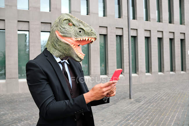 Анонімний чоловік-підприємець у гумових масках динозаврів і офіційний костюм стоячи на вулиці в місті і серфінг Інтернету на смартфоні — Stock Photo