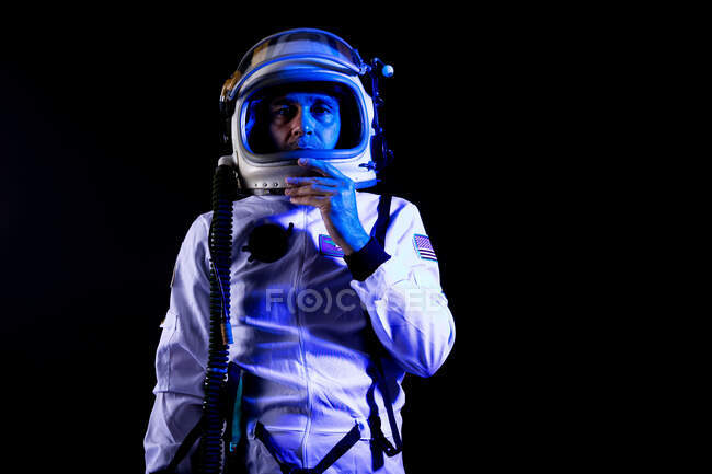Männlicher Kosmonaut mit weißem Weltraumanzug und Helm, während er auf schwarzem Hintergrund in blauem Neonlicht in die Kamera blickt — Stockfoto