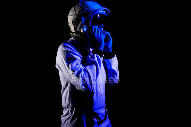 Вид сбоку на космонавта в белом скафандре и шлеме, стоящего на черном фоне в синем неоновом свете, отводящем взгляд — стоковое фото