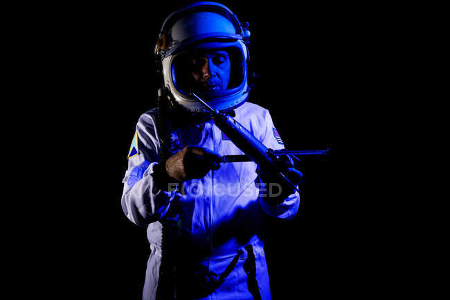 Male cosmonaut wearing white space suit and helmet while standing on black background in blue neon light holding small satellite — Stock Photo