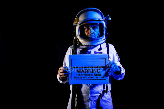 Serious adult male cosmonaut in spacesuit and helmet demonstrating placard with text Warning Restricted Area Deadly Force Authorized while standing on black background and looking at camera — Fotografia de Stock