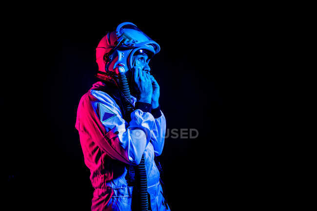 Side view of male cosmonaut wearing white space suit and helmet while standing on black background in pink and blue neon light looking away — Stock Photo
