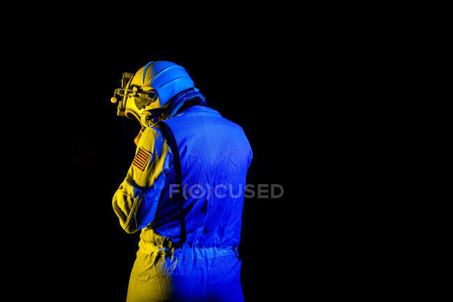 Back view of unrecognizable male cosmonaut wearing white space suit and helmet while standing on black background in blue and yellow neon light — Stock Photo