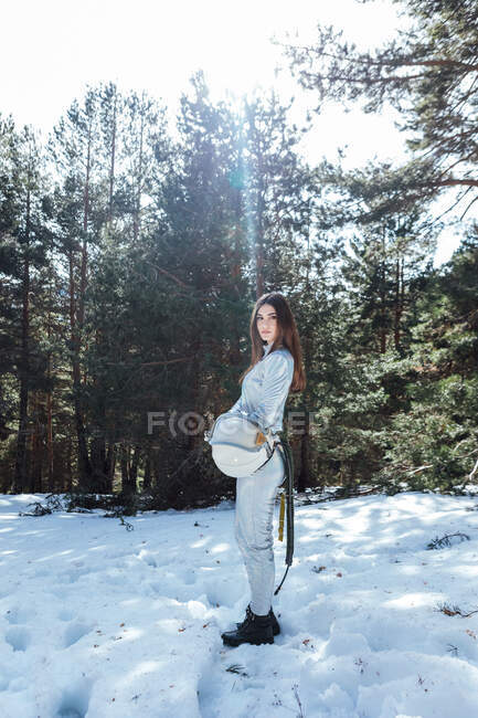 Side view of female cosmonaut in spacesuit holding helmet in hands and standing in snowy woodland while looking away — Stock Photo