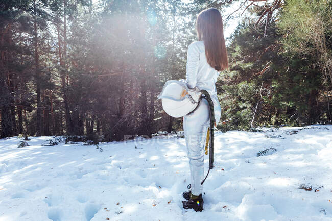 Side view of unrecognizable female cosmonaut in spacesuit holding helmet in hands and standing in snowy woodland — Stock Photo