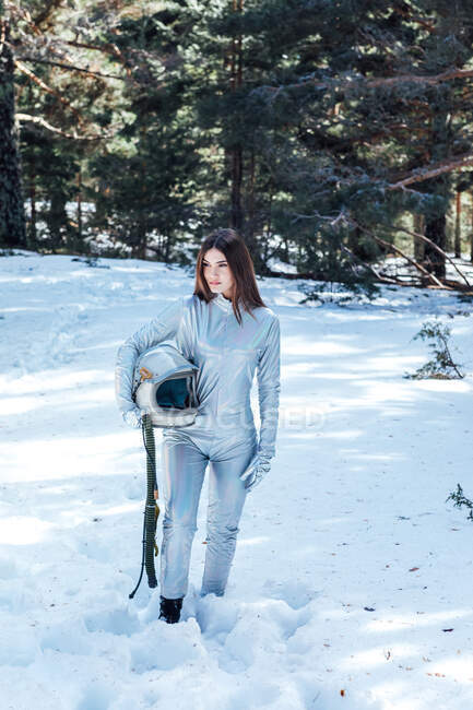 Cosmonauta fêmea em trajes espaciais segurando capacete nas mãos e em pé na floresta nevada enquanto olha para a câmera — Fotografia de Stock