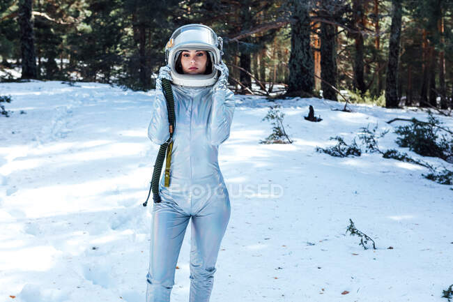 Focused young female astronaut in spacesuit and helmet looking at camera and standing in snowy woodland — Stock Photo