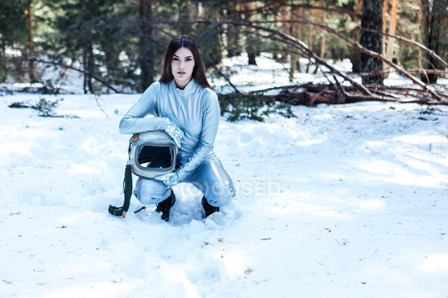Female cosmonaut in spacesuit holding helmet in hands crouched in snowy woodland while looking at camera — Photo de stock