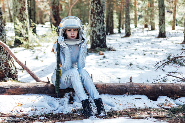 Astronauta joven enfocada en traje espacial y casco mirando a la cámara y sentada en el tronco de un árbol en un bosque nevado - foto de stock