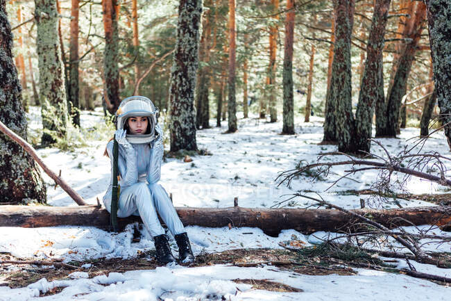 Focused young female astronaut in spacesuit and helmet looking away and sitting on tree trunk in snowy woodland — Fotografia de Stock