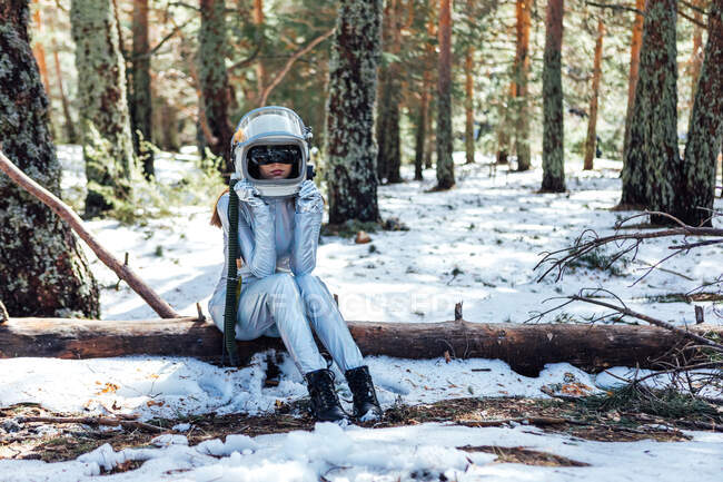 Неузнаваемая молодая женщина-астронавт в скафандре и шлеме сидит на стволе дерева в снежном лесу — стоковое фото