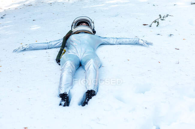 Ganzkörperfitte ruhige Raumfahrerin in Kostüm und Helm liegt mit ausgestreckten Armen auf schneebedeckter Lichtung im Winterwald — Stockfoto