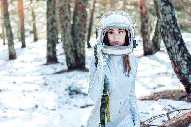 Astronauta joven enfocada en traje espacial y casco mirando hacia otro lado y de pie en un bosque nevado - foto de stock