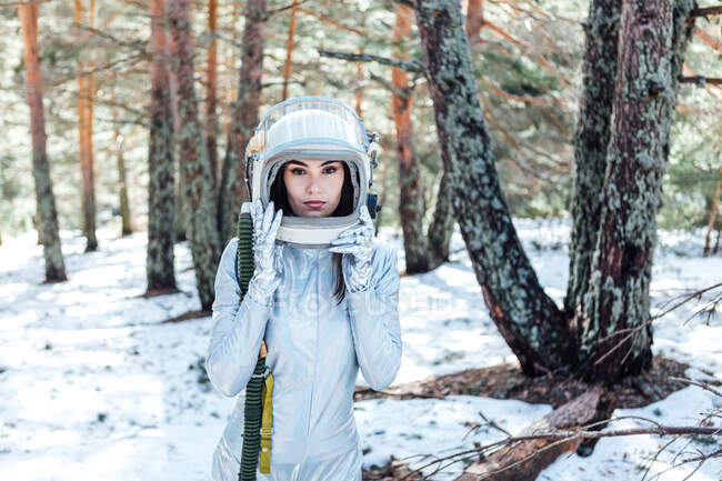 Focused young female astronaut in spacesuit and helmet looking at camera and standing in snowy woodland — Fotografia de Stock
