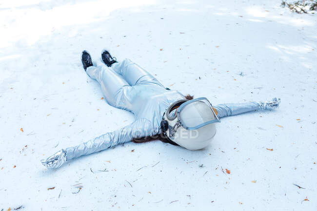 Espaciadora en forma de cuerpo completo en traje y casco tumbado con los brazos extendidos en el claro nevado en el bosque de invierno - foto de stock