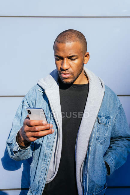 Hombre afroamericano adulto sin afeitar con la mano en el bolsillo navegando por Internet en el teléfono celular contra la pared a la luz del sol - foto de stock