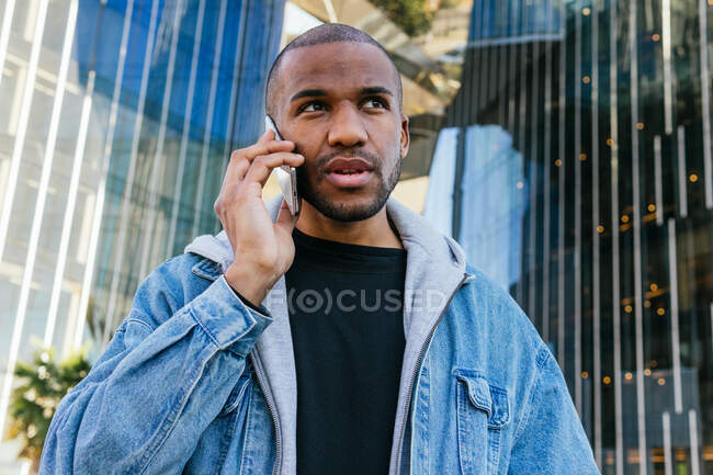 Adult bearded ethnic male in casual wear talking on mobile phone while looking away against modern urban building — Photo de stock