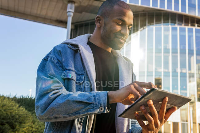 Crop content adult African American male browsing internet on tablet against contemporary building in town in sunlight — Photo de stock