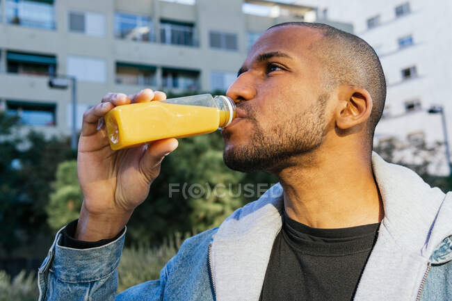 Adult bearded African American male enjoying tasty orange juice from bottle while looking forward in town — Fotografia de Stock