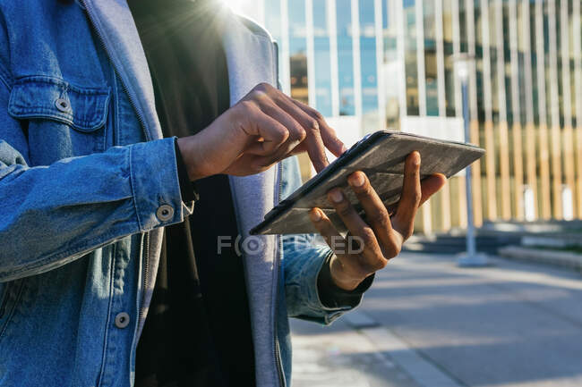 Crop unrecognizable adult African American male browsing internet on tablet against contemporary building in town in sunlight — Stock Photo