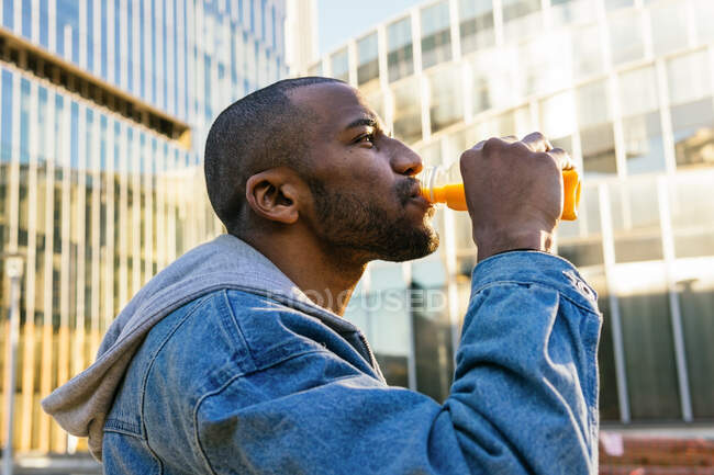 Adult bearded African American male enjoying tasty orange juice from bottle while looking forward in town — Fotografia de Stock