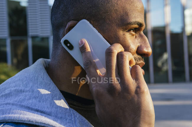 Side view of adult bearded ethnic male in casual wear talking on mobile phone while looking away against modern urban building — Stock Photo