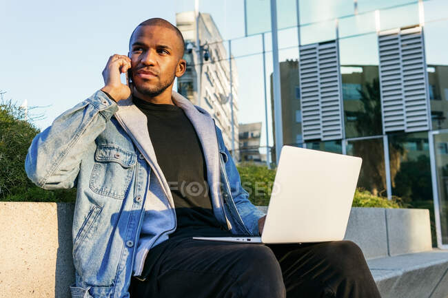 Empleado remoto masculino adulto afroamericano étnico que trabaja en la computadora portátil mientras habla por teléfono móvil sentado en la ciudad - foto de stock