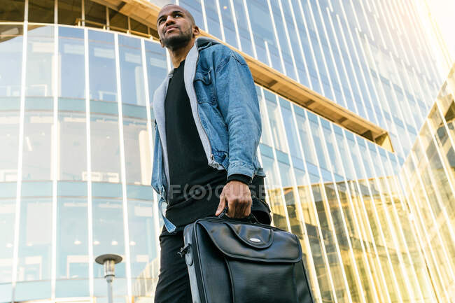 From below side view of adult ethnic male in casual outfit with leather briefcase strolling in town while looking away — Fotografia de Stock