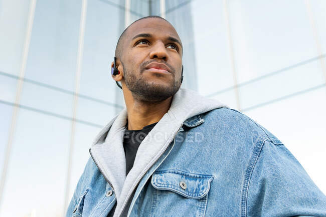 Von unten ein erwachsener bärtiger Mann mit drahtlosen Kopfhörern, der Lieder hört, während er in der Stadt wegschaut — Stockfoto
