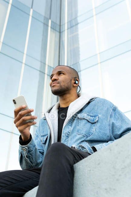 From below of adult ethnic male listening to song from earphones while sitting with cellphone in town and looking forward — Fotografia de Stock