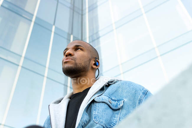 From below of adult bearded ethnic male in wireless earphones listening to song while looking away in town — Stock Photo