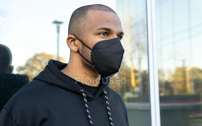 Adult bearded ethnic male in respiratory mask and black hoodie looking forward against glass wall during COVID 19 pandemic in town — Stock Photo